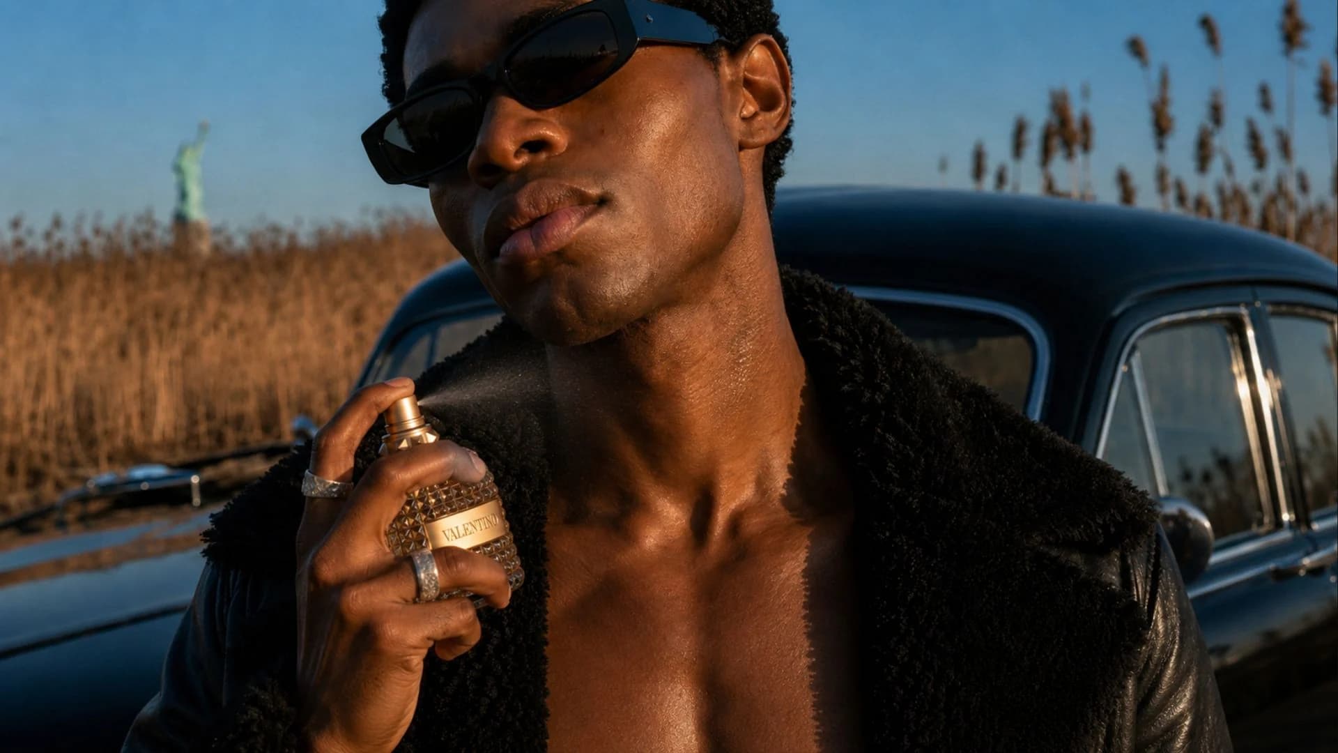 Model in sunglasses spraying Valentino Uomo Gold beside a classic car in a reed field